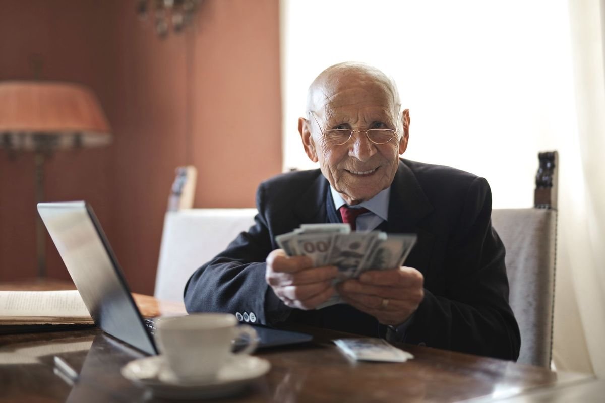 Positive senior businessman in formal suit and eyeglasses holding money in hands while sitting at table near laptop and looking at camera